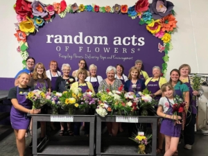 Group of women and girls standing with flower arrangements in front of purple with flower border Random Acts of Flowers sign.
